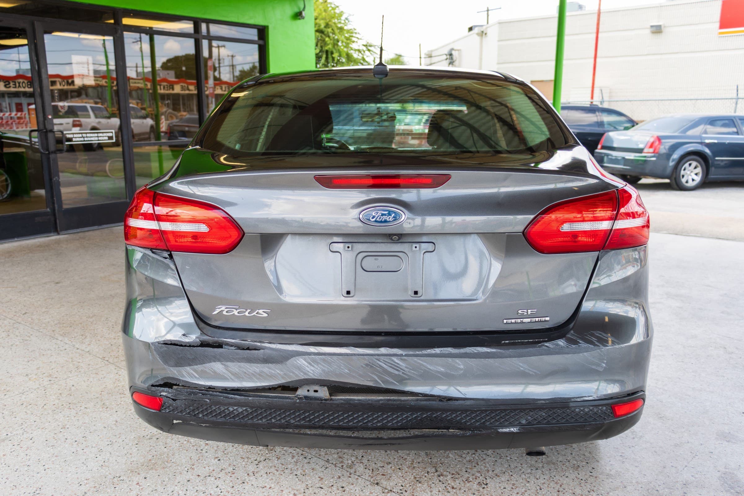 Rear view of the gray Ford Focus showing the Focus and Flex Fuel badges. The rear bumper cover is cracked and separated from the body on the driver's side, with scuffing across the bumper.