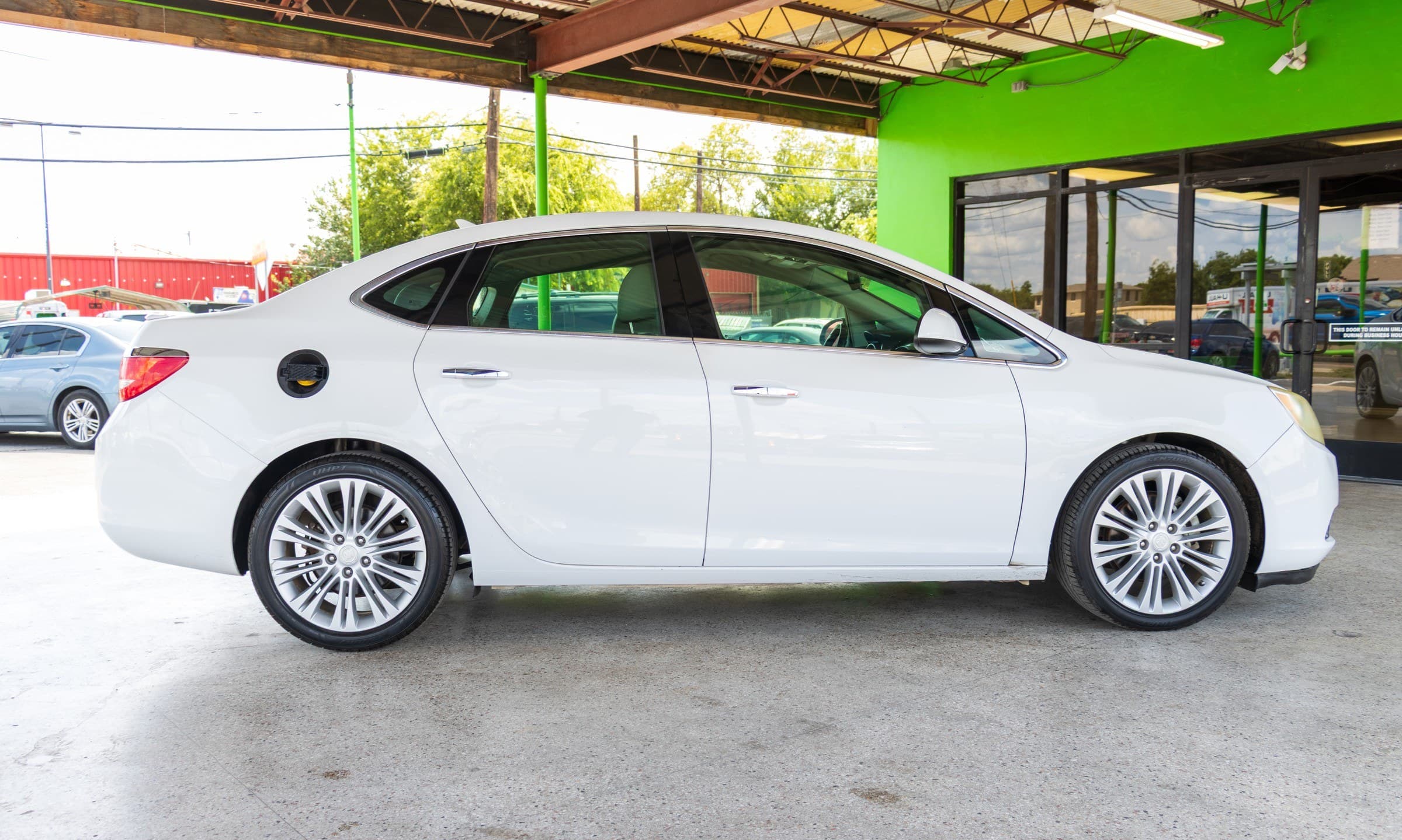 Full passenger-side profile of the white Buick Verano, showing its chrome window trim and silver multi-spoke alloy wheels.